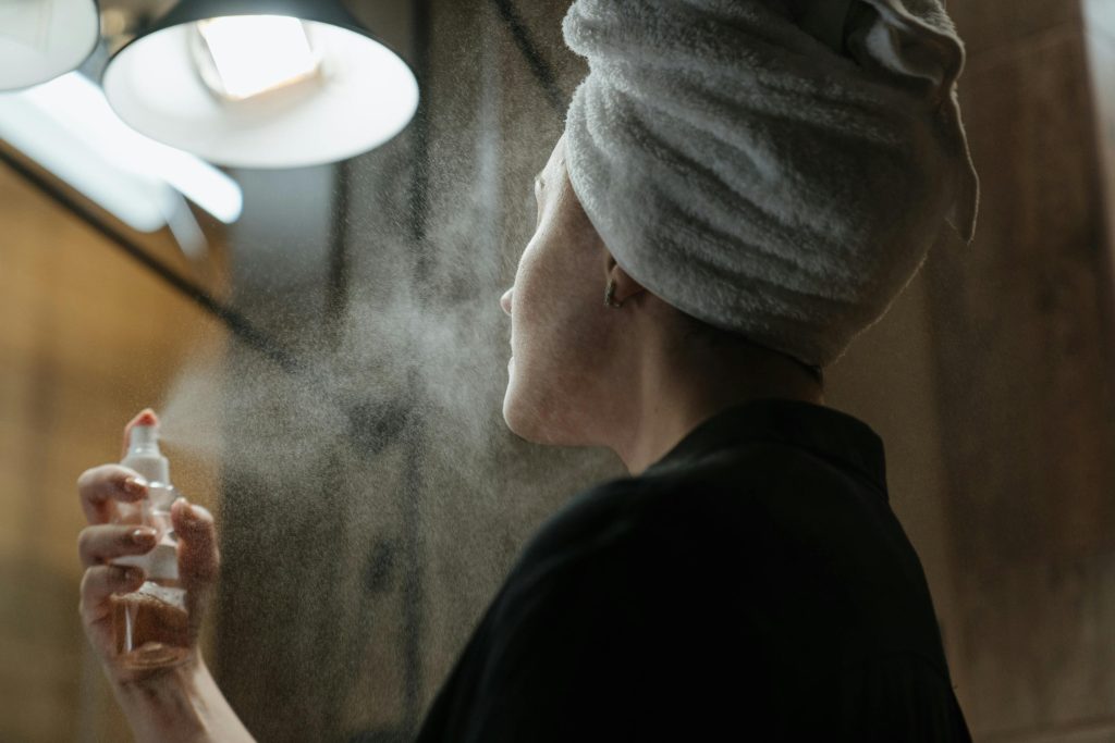 Woman misting her face with a skincare spray in a bathroom, wearing a towel wrap