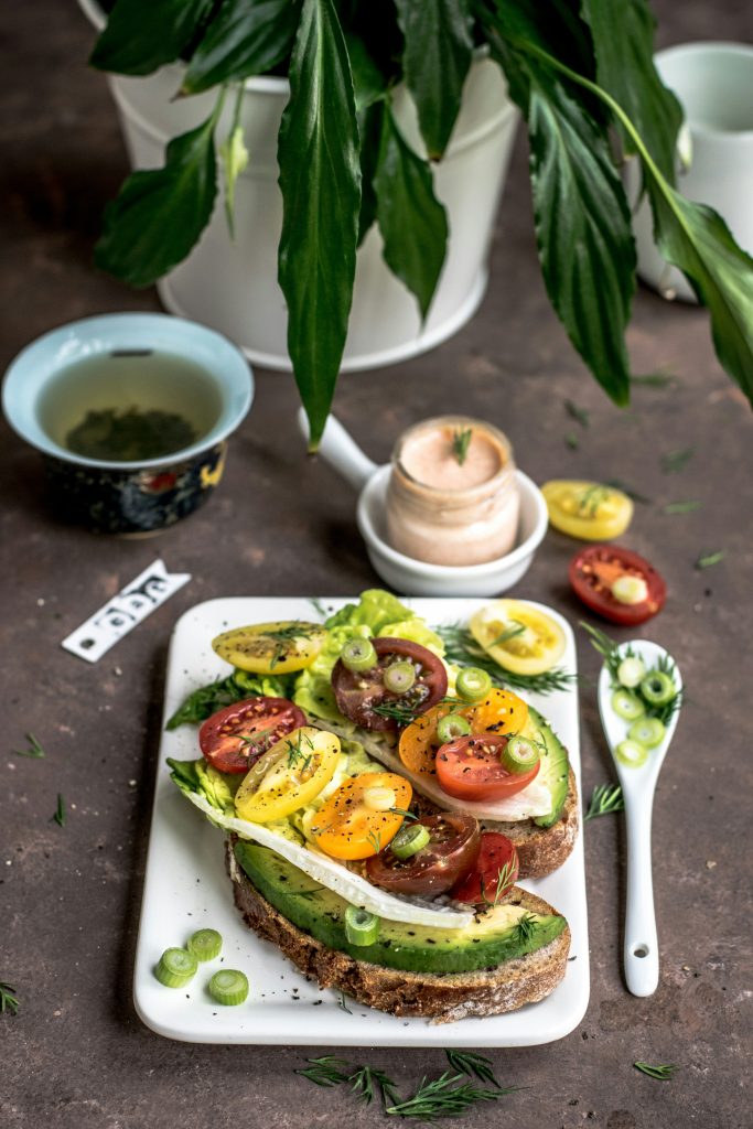 Avocado toast on whole grain bread topped with heirloom tomatoes, scallions, and fresh herbs, served with green tea.